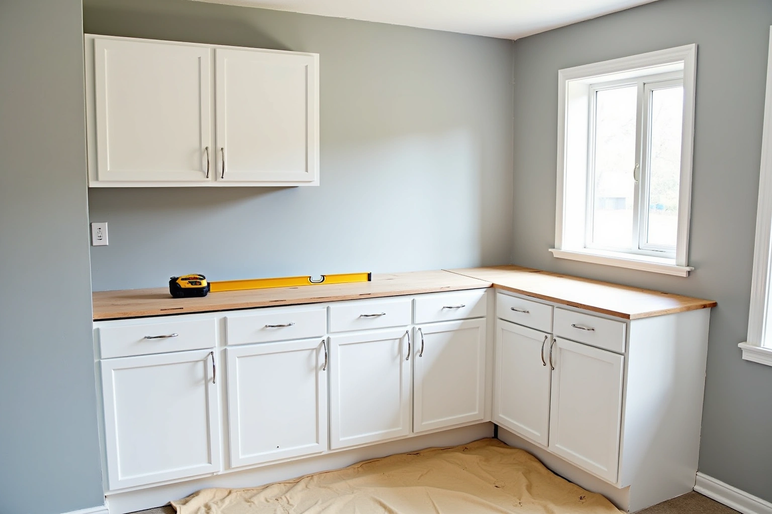 White shaker cabinets newly installed against grey walls, a bare plywood subtop holding a yellow level and drill.