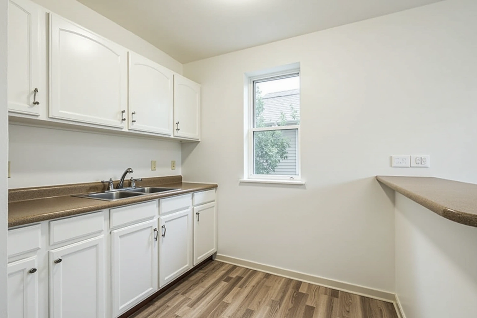 Simple white arched-panel cabinets, brown laminate counter, double sink and one small window in a bare kitchen.