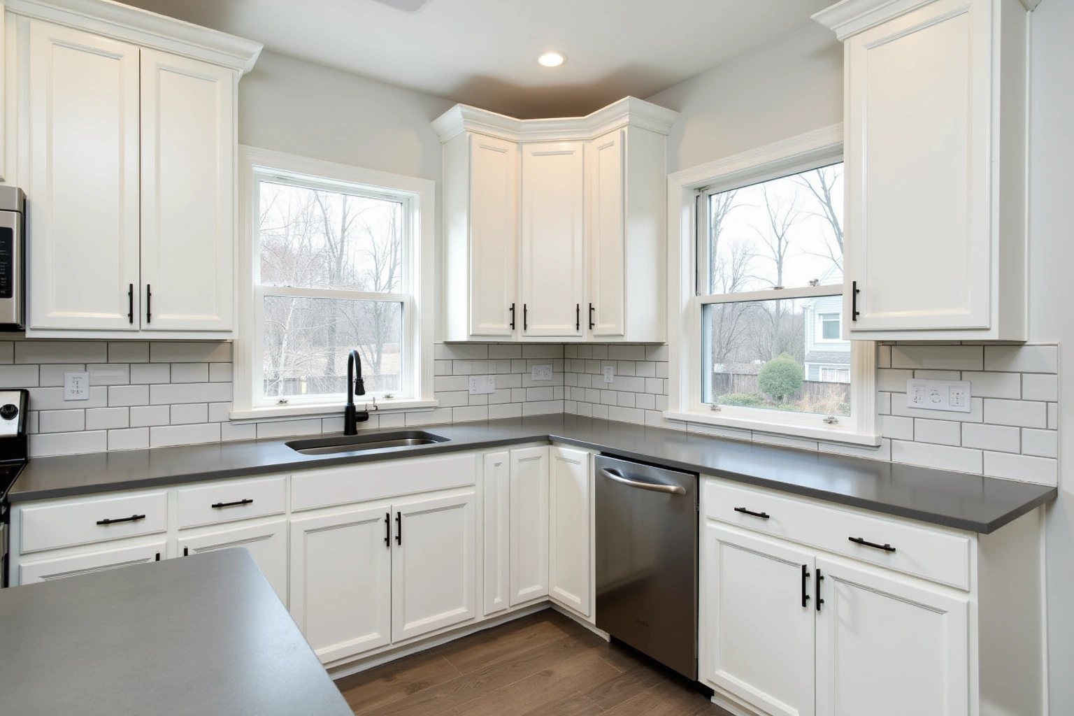 White raised-panel kitchen cabinets with matte black pulls, grey counters, white subway tile and two windows onto a yard.